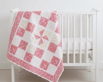 May include: A pink and white patchwork quilt with a pinwheel design, draped over a white crib. The quilt features squares of floral and solid fabrics, with a floral border. The crib has vertical slats.