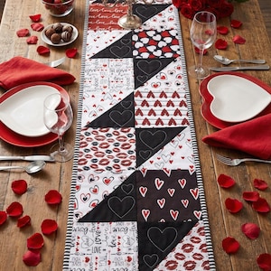 Könnte beinhalten: Eine Valentinstischdekoration mit einem gesteppten Tischläufer mit Herzen und Lippenmustern. Der Tisch ist mit herzförmigen Tellern, roten Servietten, Weingläsern und Erdbeeren gedeckt. Ein Strauß roter Rosen und Kerzen vervollständigen die Szene.