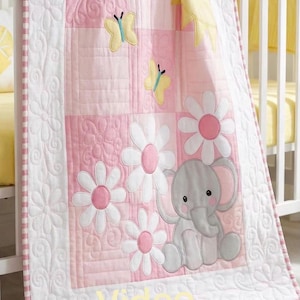Puede incluir: Una manta acolchada para bebé con un diseño de patchwork en tonos rosa y blanco. La manta muestra mariposas amarillas aplicadas, margaritas blancas con centros rosas, un sol amarillo y un elefante gris. La palabra Video está impresa en amarillo.
