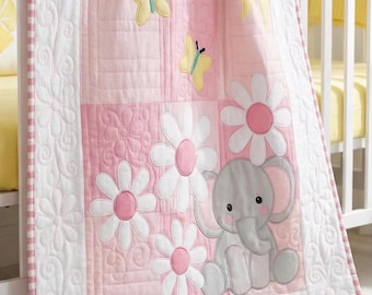 May include: A quilted baby blanket featuring a patchwork design in shades of pink and white. The blanket displays appliqued yellow butterflies, white daisies with pink centers, a yellow sun, and a gray elephant. The word Video is printed in yellow.