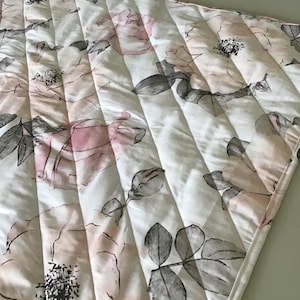 May include: A quilted fabric with a floral pattern. The design features large pink and gray roses and leaves on a white background. The fabric is stitched with vertical lines, creating a textured effect. The edges are finished with a white border.