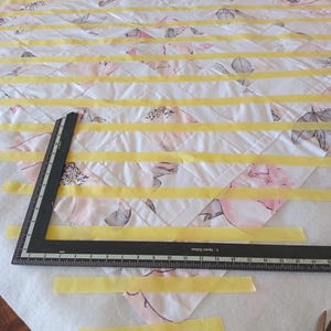 May include: A quilt in progress, featuring a white fabric with a floral pattern in pink and gray. Yellow tape is used to mark quilting lines. A black ruler is positioned on the quilt, indicating measurements.