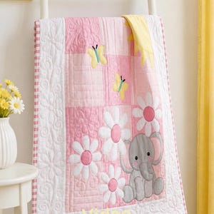 May include: A quilted baby blanket featuring a patchwork design in shades of pink and white. The blanket displays appliqued motifs of a gray elephant, yellow butterflies, a yellow sun, and white daisies with pink centers. The word Video is printed in yellow.