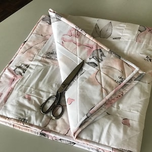 May include: A folded floral quilt with a white background and pink and gray flower pattern. A pair of silver scissors rests on the quilt. The quilt is made of quilted fabric.