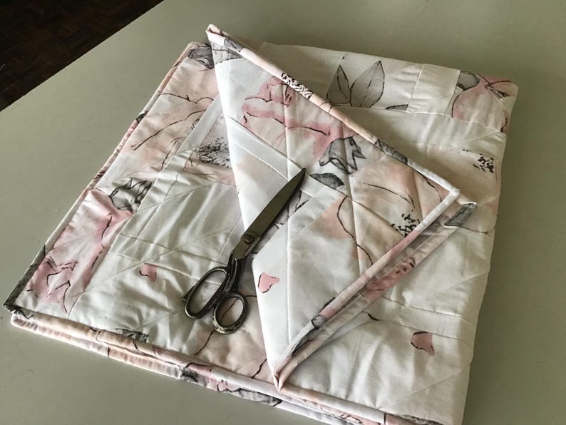 May include: A folded floral quilt with a white background and pink and gray flower pattern. A pair of silver scissors rests on the quilt. The quilt is made of quilted fabric.