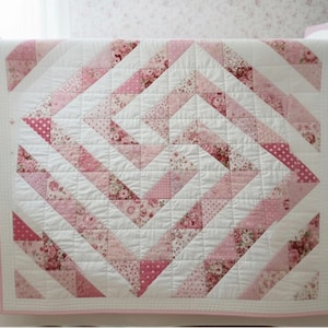 May include: A baby quilt with a geometric design in shades of pink and white. The quilt features a repeating pattern of triangles and squares, with floral and polka dot accents. The quilt is displayed on a white crib.