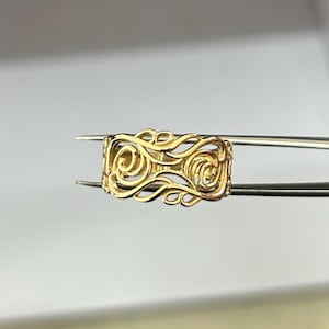 Puede incluir: Un anillo dorado con un diseño calado sostenido por unas pinzas. El anillo presenta un intrincado patrón arremolinado. El anillo tiene forma rectangular con bordes redondeados. El fondo es blanco y gris neutro.