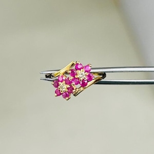 Op de afbeelding: Een gouden ring met een cluster van roze edelstenen en kleine witte edelstenen. De ring wordt vastgehouden door een pincet, wat het gedetailleerde ontwerp benadrukt. De edelstenen zijn gerangschikt in een bloemmotief.