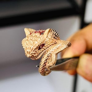 Puede incluir: Un anillo dorado con forma de serpiente con cabeza de zorro. El anillo tiene ojos de piedras preciosas rojas y lo sujeta una pinza. Una pieza de joyería única.
