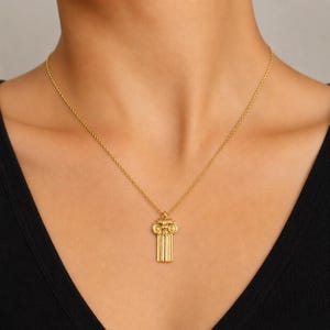 Pode incluir: Um colar de tom dourado com um pingente em forma de capitel de coluna jónica clássica com um fuste estriado. O pingente pende de uma corrente fina e delicada de tom dourado que repousa sobre o pescoço, exibindo um acabamento metálico polido.