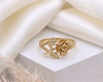 Può includere: Un anello dorato con un design dettagliato di rosa e foglie. L'anello ha una finitura strutturata e la rosa è il punto focale. L'anello è presentato su una superficie bianca con un tessuto morbido e bianco sullo sfondo.