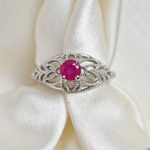 Op de afbeelding: Een zilverkleurige ring met een ronde, levendige rozerode edelsteen in het midden. De band is vervaardigd met een ingewikkeld, opengewerkt filigraanontwerp dat doet denken aan delicate, symmetrische bladvormige patronen rondom de steen.