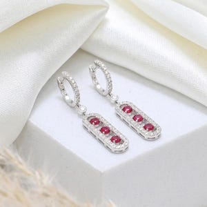 May include: Silver drop earrings with a rectangular design, each featuring three red gemstones and small, clear stones. The earrings have a hinged hoop closure. They are displayed on a white surface, showcasing their elegant design.