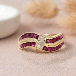 May include: A gold ring with rows of square-cut, deep red gemstones and a central section of baguette-cut, clear gemstones. The ring has a curved design, set against a soft pink background.