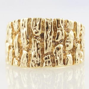 May include: A gold-colored ring with a textured surface, resembling tree bark. The ring has a wide band and a reflective surface, showcasing the intricate details of the design. This piece of jewelry is a statement ring.