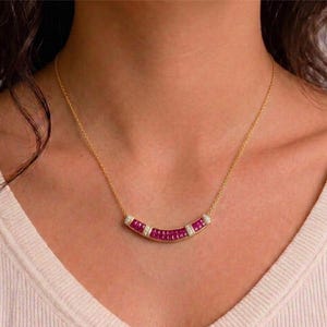 Peut inclure: Un collier en or avec un pendentif en forme de barre incurvée. Le pendentif est serti d'une rangée de petites pierres précieuses rectangulaires rouge foncé, entrecoupées de sections de petites pierres précieuses claires. Le collier est porté autour du cou.