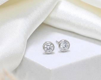 May include: A pair of silver stud earrings with round, clear gemstones. The earrings are set in a bezel setting and are displayed on a white surface with a white, silky fabric in the background. The gemstones sparkle.