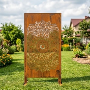 May include: A rectangular, weathered metal garden screen with a large, intricate mandala design cut into the surface. The screen is a rusty brown color and stands on two legs, set in a grassy yard with a house and trees in the background.