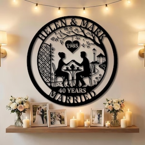 Könnte beinhalten: Ein schwarzes Metall-Wandbild mit den Namen "HELEN & MARK" und dem Jahr "1985" in einem Kreis. Das Kunstwerk zeigt ein Paar an einem Tisch, darunter die Worte "40 YEARS MARRIED". Das Bild wird über einem Regal mit Fotos und Kerzen präsentiert.