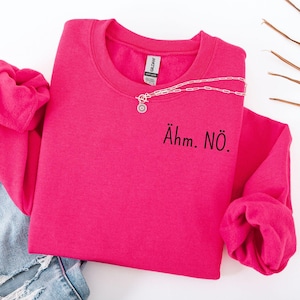 Könnte beinhalten: Ein leuchtend pinkfarbenes Sweatshirt mit dem Text "Ähm. NÖ." in Schwarz. Eine silberne Halskette mit Anhänger ist über dem Sweatshirt drapiert. Hellblaue Jeans sind auf der linken Seite des Bildes zu sehen.