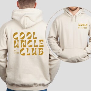 Könnte beinhalten: Cremefarbener Kapuzenpullover mit Fronttasche und Kapuze. Auf der Rückseite des Hoodies steht in Senfgelb "COOL UNCLE CLUB EST. 2025". Auf der Vorderseite steht das Wort "UNCLE EST. 2025" in der gleichen Senffarbe.