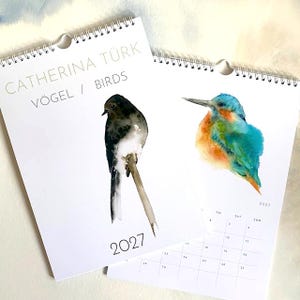 Könnte beinhalten: Zwei Wandkalender für 2027 mit weißen Seiten und Spiralbindung. Ein Deckblatt zeigt ein Aquarell eines schwarz-weißen Vogels auf einem Ast mit dem Text CATHERINA TÜRK VÖGEL / BIRDS. Das andere zeigt einen farbenfrohen Eisvogel.