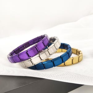 May include: Three colorful bracelets in purple, silver, blue, and gold. Each bracelet is made of rectangular links, creating a flexible, connected band. The bracelets are displayed on a white, textured surface.