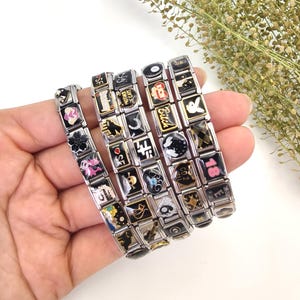 May include: Several stainless steel charm bracelets with black and gold enamel charms. The charms feature various designs, including flowers, symbols, and text. The bracelets are displayed against a white background.