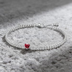 May include: A silver bracelet with a heart-shaped, deep red gemstone at the center. The bracelet is adorned with small, clear gemstones and has a clasp closure. The jewelry is displayed on a gray, textured surface.
