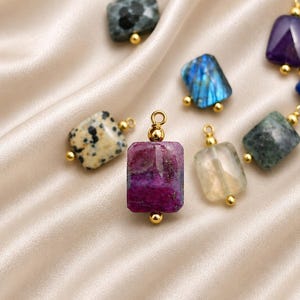 May include: A collection of gemstone pendants in various colors, including purple, blue, green, and speckled beige. Each rectangular pendant has a gold-colored bail and a small gold bead at the bottom. The pendants are displayed on a satin fabric.
