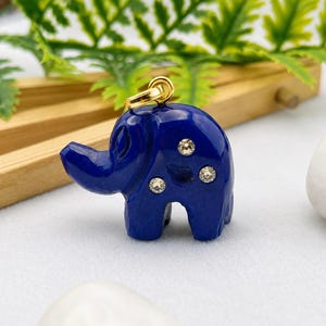 Puede incluir: Un colgante con forma de elefante de color azul intenso que presenta tres pequeñas piedras redondas y transparentes incrustadas en el lateral. El colgante incluye una anilla de metal dorado en la parte superior para sujetarlo a una cadena.