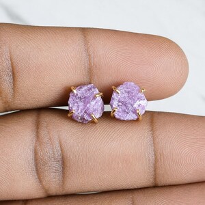 May include: A pair of stud earrings featuring raw, light purple gemstones set in gold-toned metal prongs. Each stone has an irregular, textured surface and a faceted appearance, securely held by four small metal claws on each earring base.