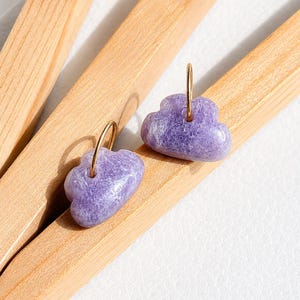 Pode incluir: Um par de brincos de argola com pingentes roxos em forma de nuvem com uma superfície texturizada e cristalina. Cada pingente está suspenso por uma fina argola de metal dourado, criando um design simples que destaca a pedra roxa natural.