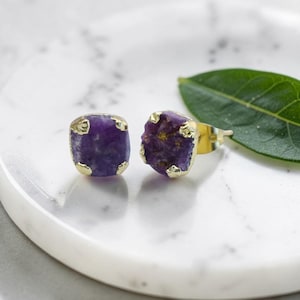 Peut inclure: Une paire de boucles d'oreilles carrées avec des pierres précieuses violettes taillées à l'état brut et des bordures en métal doré. Les boucles d'oreilles sont présentées sur une surface en marbre blanc. Les boucles d'oreilles sont un bijou.