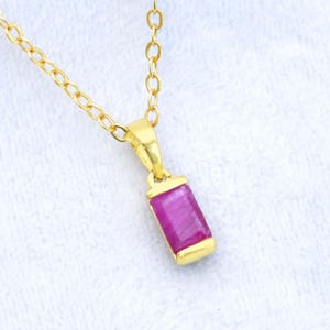 May include: A gold-colored necklace with a rectangular, magenta-colored pendant. The pendant is set in a gold bezel and hangs from a delicate gold chain. The background is a soft, white fabric.