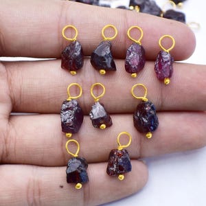 Può includere: Una collezione di pendenti in granato rosso scuro grezzo, con anelli dorati e piccole perline dorate. I pendenti, di forma irregolare, sembrano pietre preziose naturali, ideali per la creazione di gioielli.