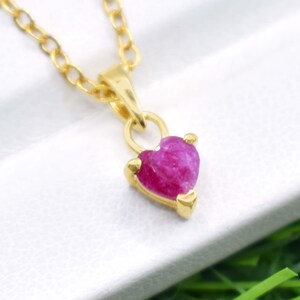 Peut inclure: Un collier doré avec un pendentif en forme de cœur en pierre précieuse rose. Le pendentif est d'un rose vif et est serti dans un métal doré. La chaîne est également dorée et présente un design à maillons délicats. Le collier est présenté sur une surface blanche.