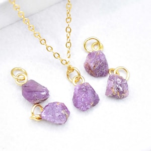 May include: A gold-coloured chain necklace with a raw, purple gemstone pendant. Several loose gemstones with gold-coloured loops are scattered around the necklace. The gemstones have a rough, natural texture and a light purple hue.
