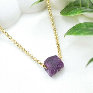 May include: A gold-colored chain necklace featuring a raw, dark purple gemstone pendant. The necklace is displayed on a white surface with green foliage in the background. The gemstone has a rough, natural texture.