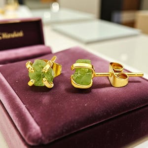 Puede incluir: Un conjunto de anillo y pendientes de oro con piedras preciosas de color verde claro en bruto. El anillo tiene una banda de oro con garras que sujetan la piedra. El pendiente es un pendiente de botón con una piedra preciosa y un engaste de oro similares. La joyería se exhibe sobre una superficie de terciopelo púrpura.