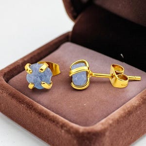 Puede incluir: Un anillo y un par de pendientes dorados, cada uno con una piedra preciosa azul claro en bruto. Los pendientes son de tipo botón. Las joyas se presentan en un joyero de terciopelo marrón.