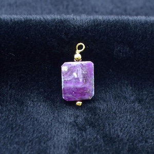 Puede incluir: Un colgante rectangular de piedra preciosa púrpura con detalles dorados. El colgante tiene un lazo dorado en la parte superior y una pequeña cuenta dorada en la parte inferior. La piedra preciosa es de color púrpura intenso con algunas variaciones más claras.