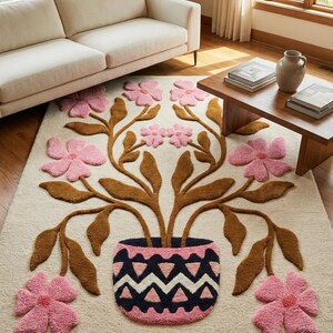 May include: A cream-coloured rug with a floral design. The rug features a central vase with brown stems and pink flowers. Additional pink flowers are scattered around the vase. The vase has a navy blue and pink zig-zag pattern.