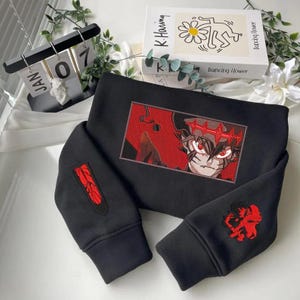 Puede incluir: Sudadera negra con diseño de personaje de anime bordado en rojo. El diseño está enmarcado en un rectángulo rojo. Bordado rojo adicional en las mangas. La sudadera se muestra sobre una superficie blanca.