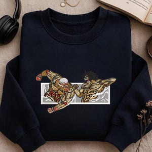May include: Navy blue sweatshirt with an embroidered design of two muscular figures in a fighting pose. The figures are in shades of tan, red, and white, set against a gray and white rectangular frame. The sweatshirt has a crew neck.