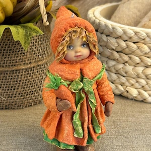 Op de afbeelding: Handgemaakte pop gekleed in een oranje outfit die op een wortel lijkt. De pop heeft een capuchon, groene accenten en houdt een wortel vast. Het gezicht van de pop heeft blauwe ogen en een serene uitdrukking. Een uniek decoratief item.