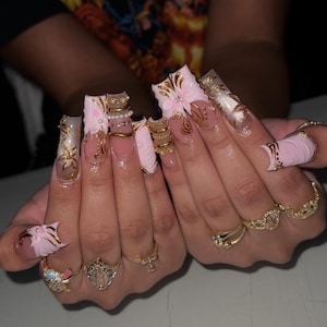 Könnte beinhalten: Nahaufnahme von langen, quadratischen Acrylnägeln. Die Nägel sind hellrosa lackiert und mit goldenen Akzenten, floralen Designs und kleinen Perlen verziert. Mehrere goldene Ringe sind auf den Fingern zu sehen.