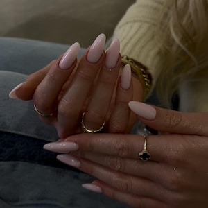 May include: Hands with long, almond-shaped, pale pink nails. The nails have a glossy finish. The hands are decorated with gold rings, including a plain band, a textured ring, and a ring with a black clover design.