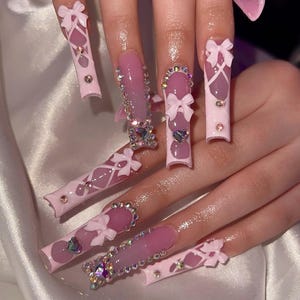 May include: Long, pink acrylic nails with various designs. Some nails feature white bows, rhinestones, and heart-shaped gems. Others have a lattice design with small gems. The nails are a light pink color with a glossy finish.