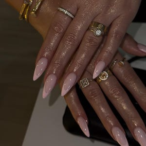 May include: Close-up of hands with long, almond-shaped nails painted in a light pink with a white tip. The hands are adorned with multiple gold rings and a gold bracelet. The nails have a glossy finish.